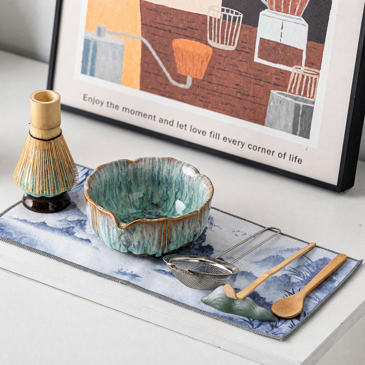 Flat lay of the matcha set on a patterned cloth: kiln-glazed bowl, bamboo whisk on its stand, a metal strainer and wooden utensils