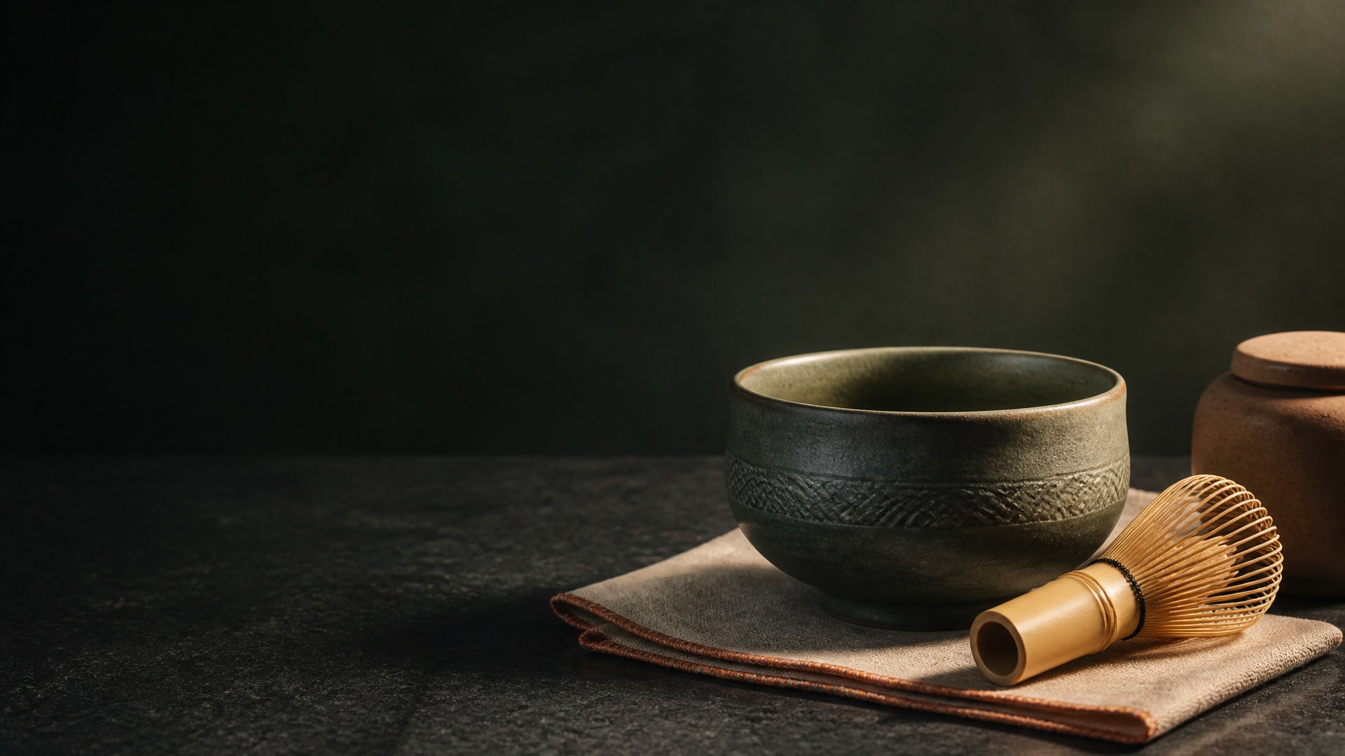 Ceramic chawan and bamboo chasen resting on a linen cloth, lit from the side